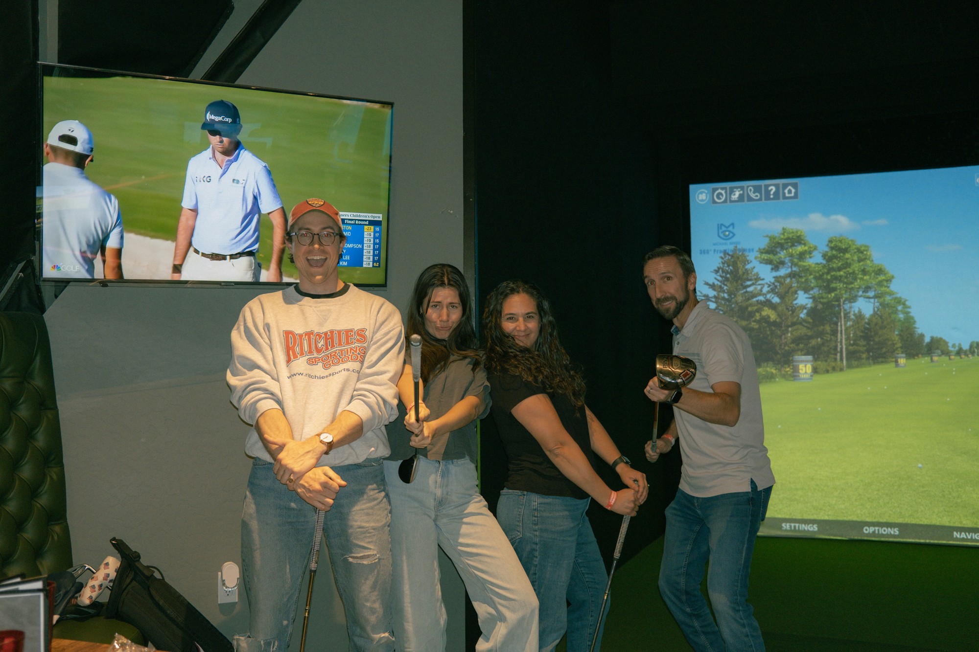 Group of four friends posing happily in front of full-screen indoor golf simulator screen holding clubs